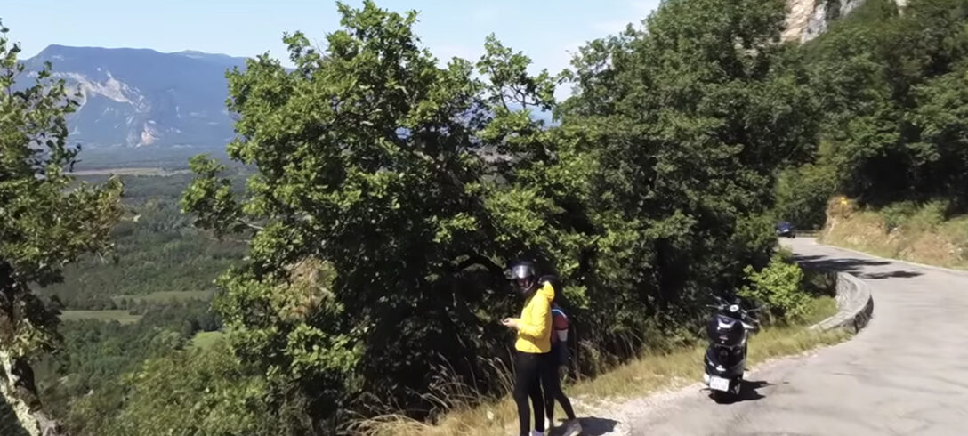 couple voyageant en scooter debout sur le côté d'une route de montagne pour prendre une photo du paysage