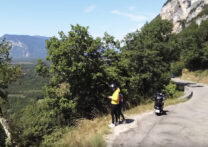 couple voyageant en scooter debout sur le côté d'une route de montagne pour prendre une photo du paysage
