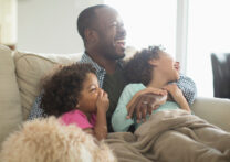 père et deux filles s'embrassant, allongés sur le canapé et recouverts d'une couverture
