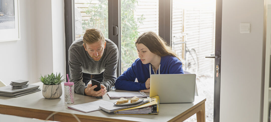 père penché sur le bureau de sa fille expliquant quelque chose sur son téléphone portable