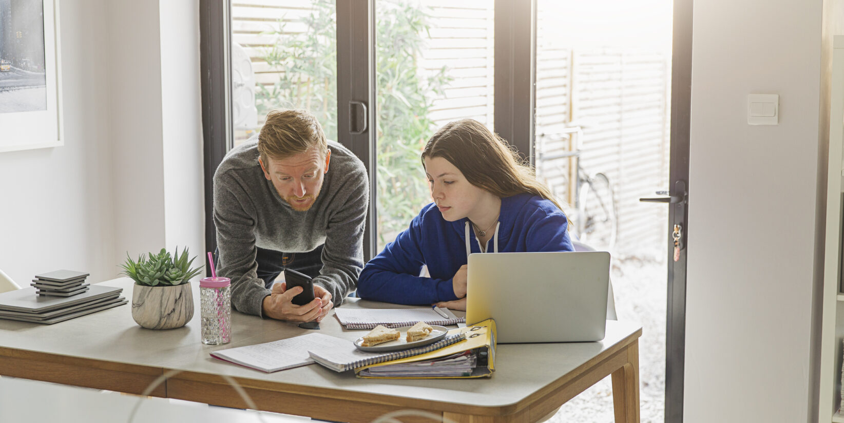 père penché sur le bureau de sa fille expliquant quelque chose sur son téléphone portable