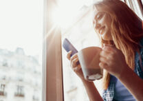 femme à la fenêtre de la maison buvant du café tout en envoyant des SMS