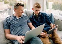 Père souriant avec un ordinateur portable regardant son fils à l'aide d'une tablette numérique assis sur un canapé à la maison