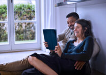couple assis sur le sol d'une pièce de la maison en riant en regardant quelque chose sur la tablette