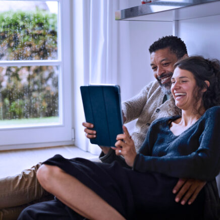 couple assis sur le sol d'une pièce de la maison en riant en regardant quelque chose sur la tablette