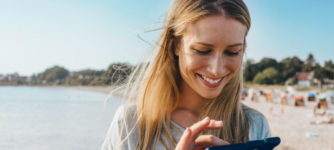 gros plan d'une jeune femme souriante utilisant son téléphone portable avec la plage en arrière-plan