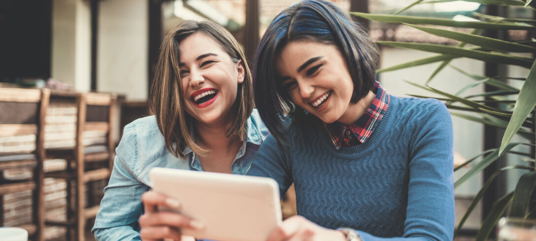 Deux jeunes femmes tenant une tablette, discutant et souriant dans un café.