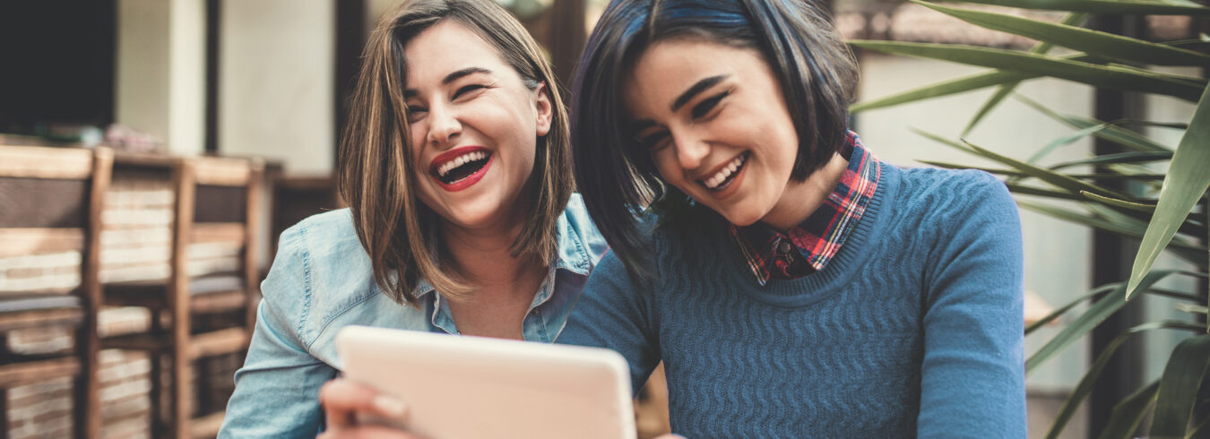 Deux jeunes femmes tenant une tablette, discutant et souriant dans un café.