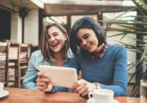 Deux jeunes femmes tenant une tablette, discutant et souriant dans un café.