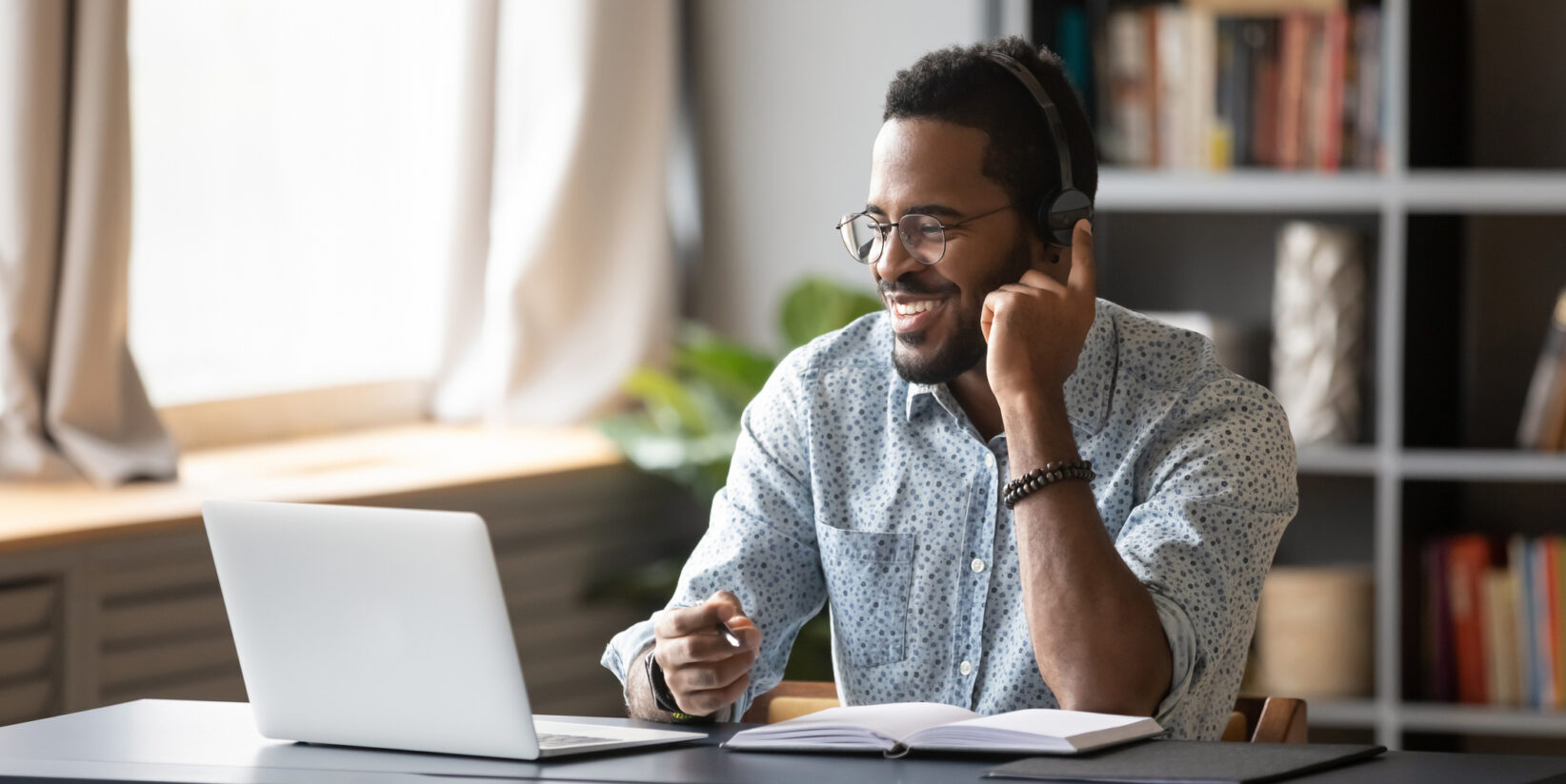 Un étudiant africain apprend à utiliser des écouteurs en ligne avec un micro et un ordinateur tient un stylo prend des notes dans un cahier assis au bureau à la maison.