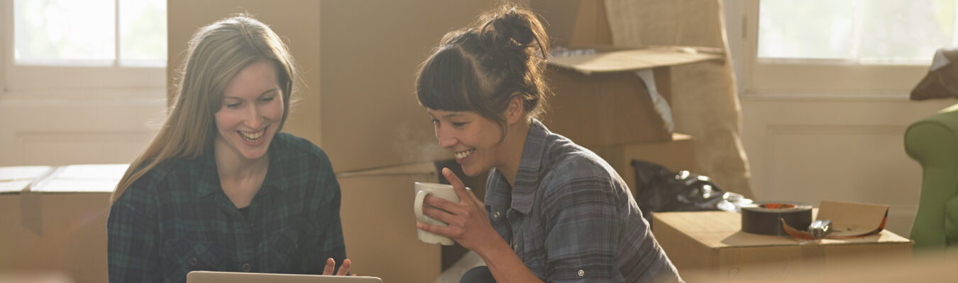 Deux jeunes femmes professionnelles assises dans un nouvel appartement entouré de boîtes d'emballage, elles boivent du thé et regardent un ordinateur portable.