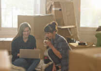 Deux jeunes femmes professionnelles assises dans un nouvel appartement entouré de boîtes d'emballage, elles boivent du thé et regardent un ordinateur portable.