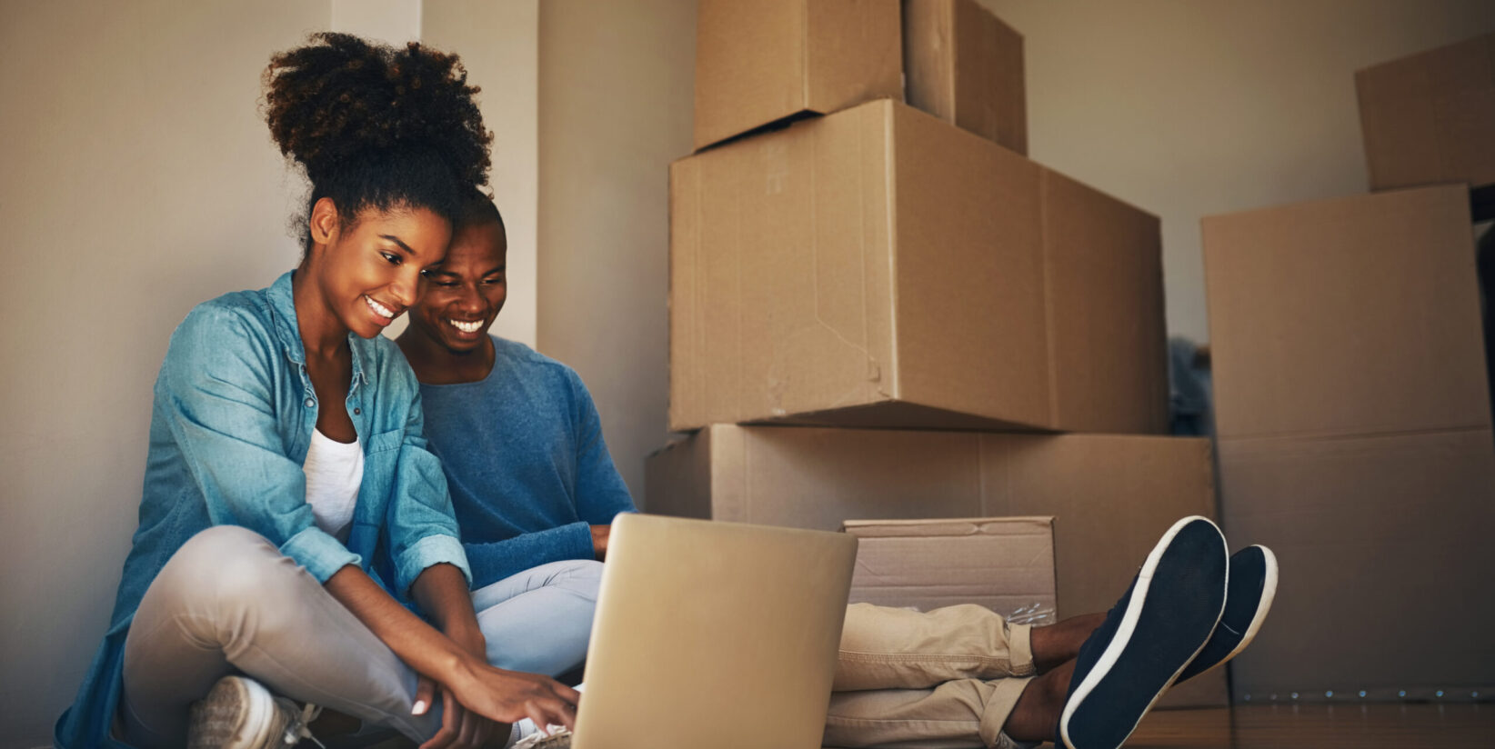 Photo d'un jeune couple joyeux naviguant ensemble sur un ordinateur portable tout en étant entouré de boîtes en carton à l'intérieur de la maison